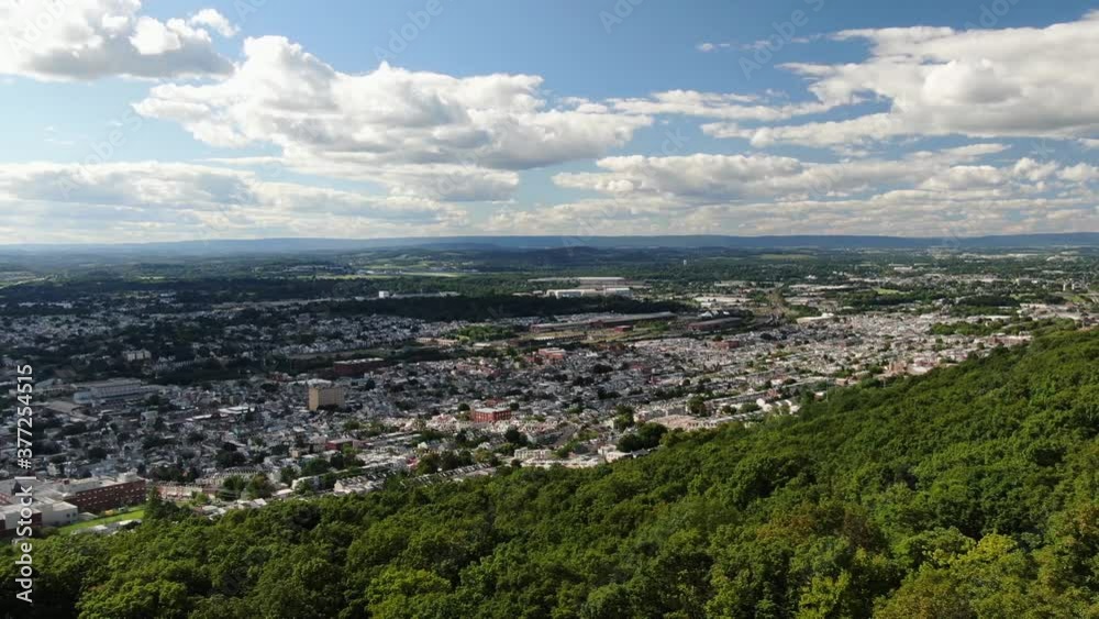 Wide angle aerial pan of densely populated urban American city, Reading Pennsylvania USA below green treetops of mountain on summer day, establishing shot Wide angle aerial pan of densely populated urban American city, Reading Pennsylvania USA below green treetops of mountain on summer day, establishing shot