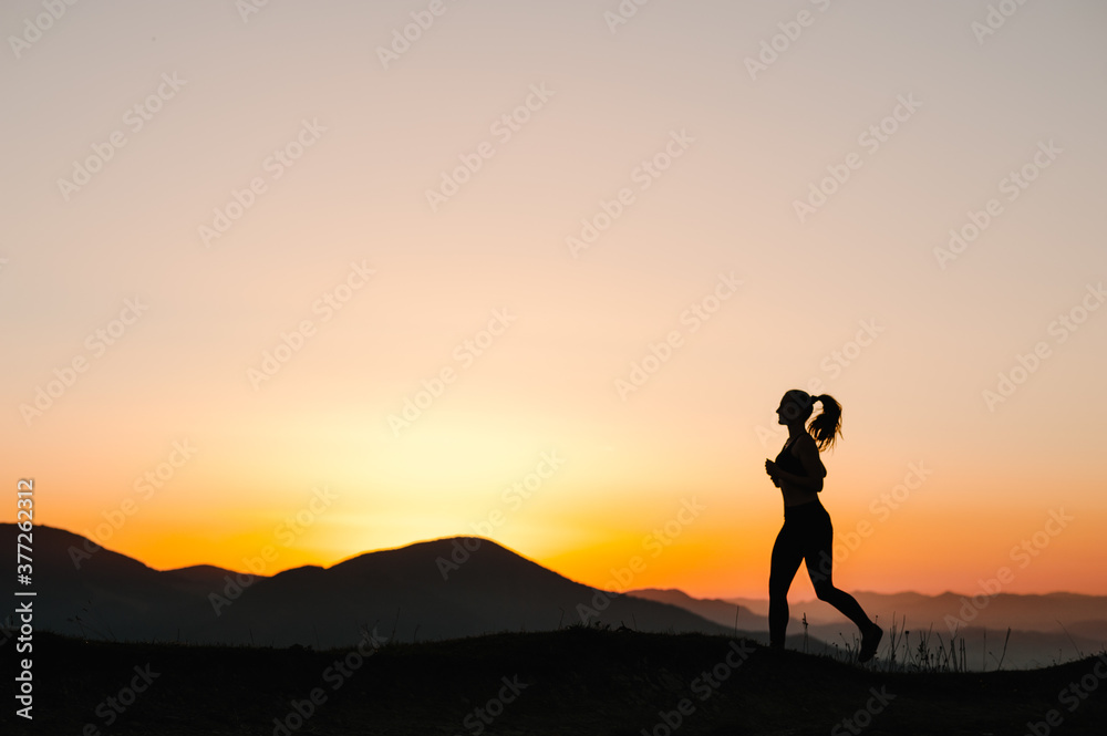 Athlete woman running on the road in the mountains. Silhouette against the sky at sunrise while running. Runner fitness girl in sport tight clothes. Bright sunset and blurry background. Horizontal.