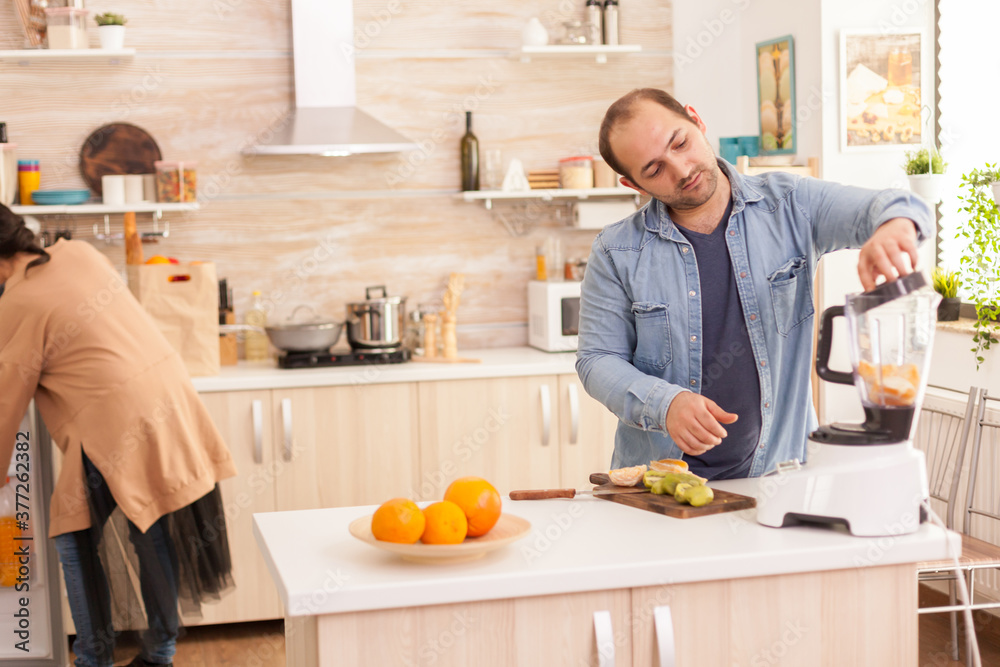 Man mixing fruits in blender while wife is looking inside refrigerator. Healthy carefree and cheerful lifestyle, eating diet and preparing breakfast in cozy sunny morning