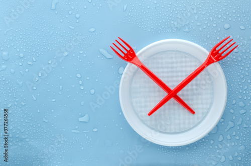 Top view of red plastic forks, white dish on the wet blue surface.Empty space.Dish and Forks as a symbol of clock and time for reducing pollution