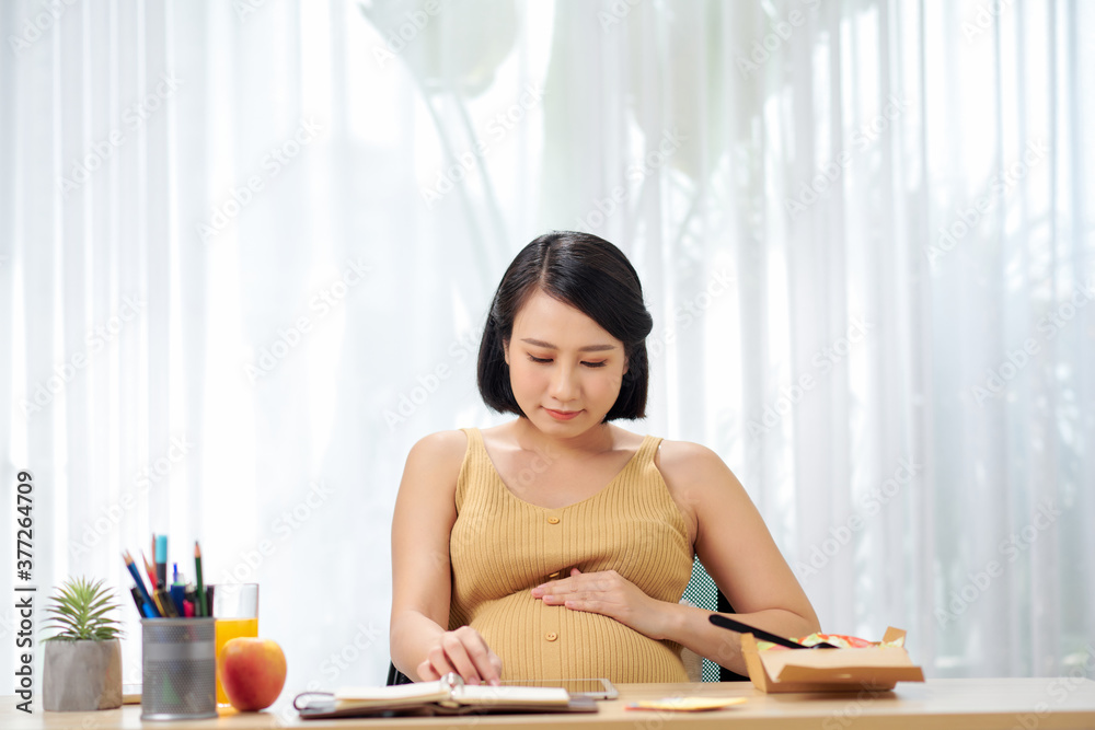 pregnant young woman sitting and using digital tablet on table in living room and have a breakfast and working pregnant young woman sitting and using digital tablet on table in living room and have a breakfast and working