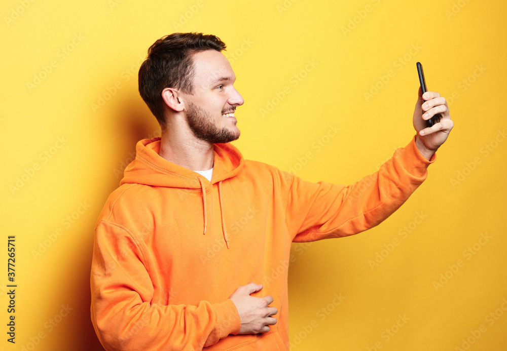 young handsome man holding mobile phone and making photo of himself while standing against yellow background young handsome man holding mobile phone and making photo of himself while standing against yellow background