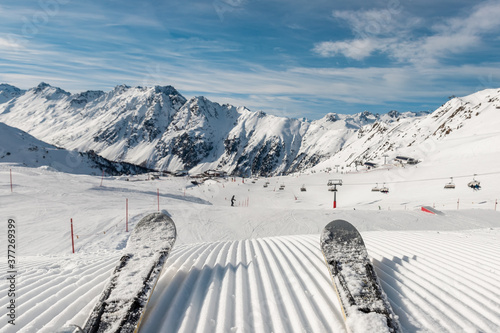 Panorama point of view skier legs on downhill start straight line rows freshly prepared groomed ski slope piste on bright day blue sky background. Snowcapped mountain landscape europe winter resort photo