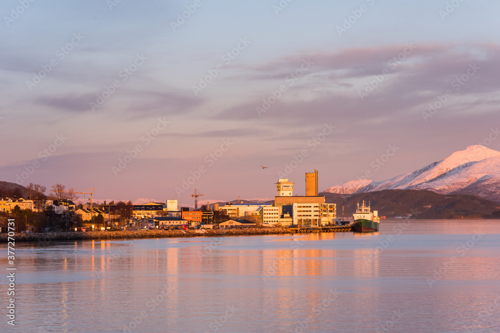 The port of the town Molde in Norway in golden evening light with snow covered mountains and starting cargo airplane in background
