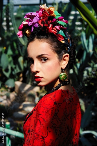 Tropical garden. A beautiful brunette with a golden tan poses in a red dress and floral headdress in green leaves. Face with clean skin, red lips.