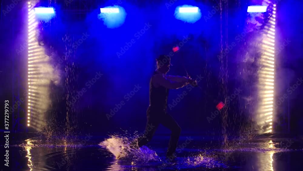 A man is silhouetted with glowing red grenade in a dark smoky studio. A stylish entertainer performs a fire show in the rain against a backdrop of backlit blue smoke. Slow motion. A man is silhouetted with glowing red grenade in a dark smoky studio. A stylish entertainer performs a fire show in the rain against a backdrop of backlit blue smoke. Slow motion.