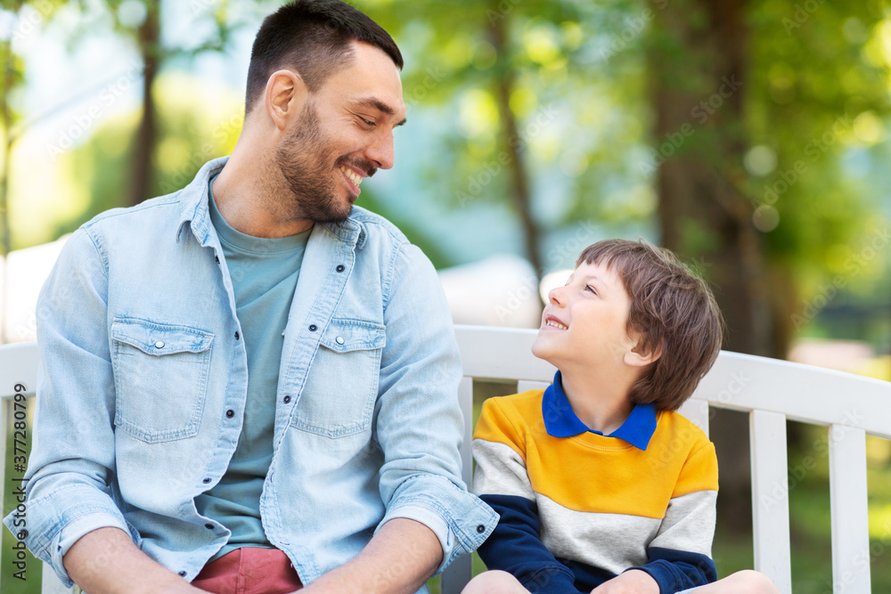 family, fatherhood and people concept - happy father and son sitting on bench at summer park and talking family, fatherhood and people concept - happy father and son sitting on bench at summer park and talking