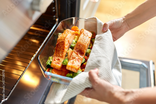 food cooking, culinary and people concept - young woman with towel taking baking dish with salmon fish and vegetables out of oven at home kitchen