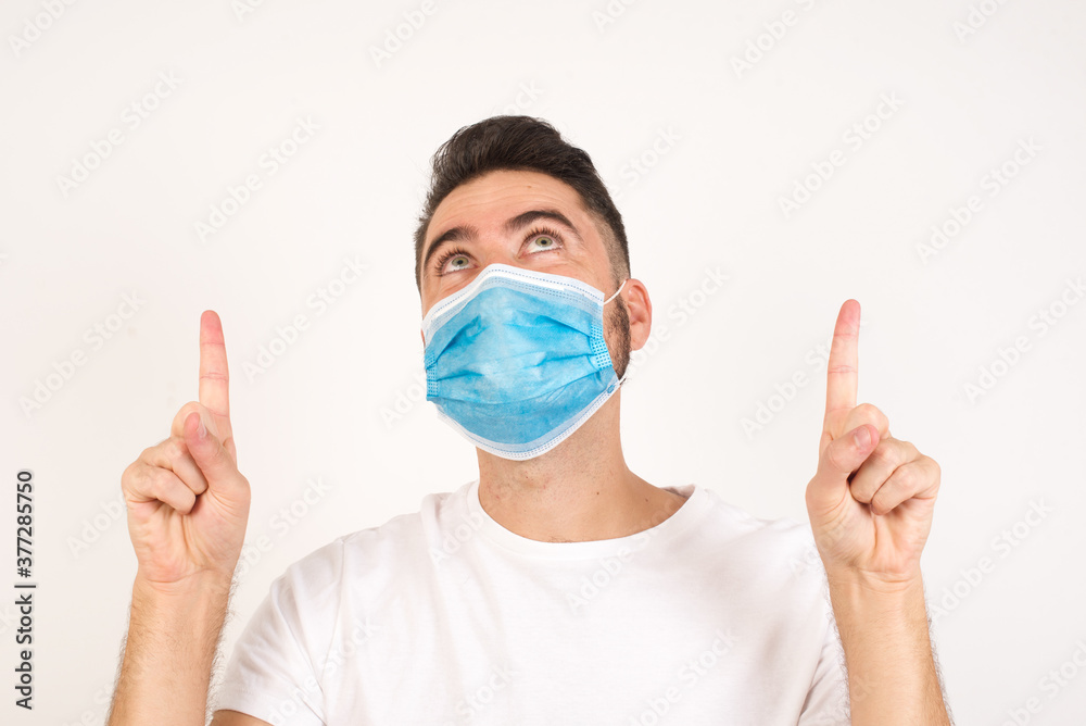 Horizontal shot of successful friendly looking young dark haired man exclaiming excitedly, pointing both index fingers up, indicating something shocking on blank copy space wall.