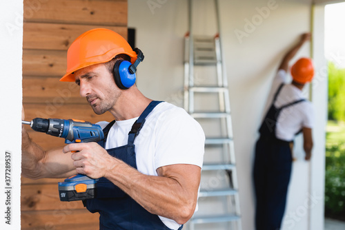 Selective focus of handyman in hardhat and uniform using electric screwdriver on facade of building