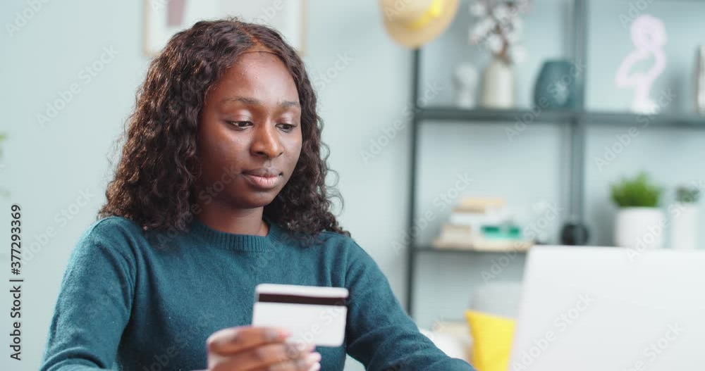 Close up portrait of cheerful African American beautiful young woman buying online on laptop. Joyful girl sitting at table in cozy room and paying with credit card on computer. Shopping concept Close up portrait of cheerful African American beautiful young woman buying online on laptop. Joyful girl sitting at table in cozy room and paying with credit card on computer. Shopping concept