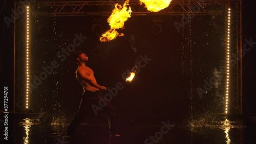 The fakir breathes fire in a dark studio in the rain. A silhouette of a half naked man looms against the background of fire in slow motion.