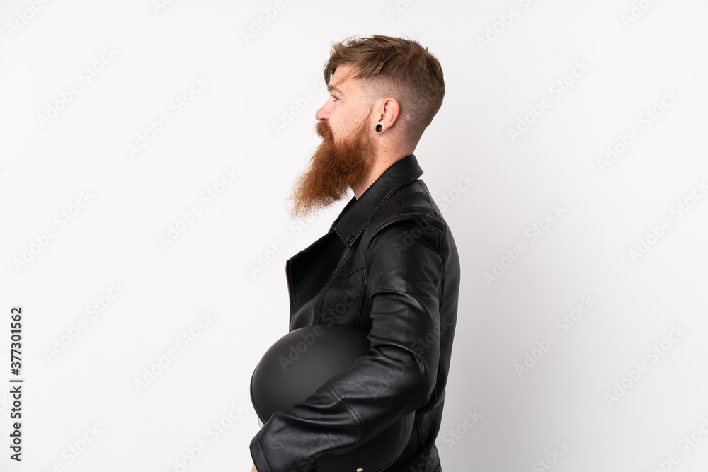 Redhead man with long beard holding a motorcycle helmet over isolated white background looking to the side Redhead man with long beard holding a motorcycle helmet over isolated white background looking to the side