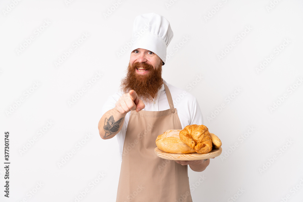Redhead man in chef uniform. Male baker holding a table with several breads points finger at you with a confident expression