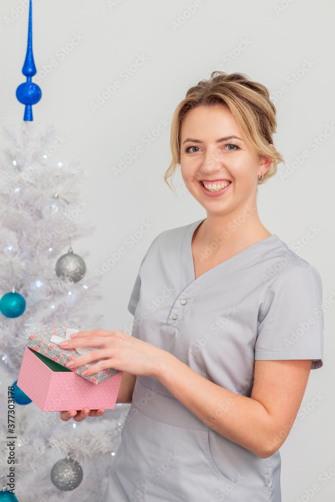 Beautiful young caucasian woman smiling in medical uniform with gift box near christmas tree on light background Beautiful young caucasian woman smiling in medical uniform with gift box near christmas tree on light background
