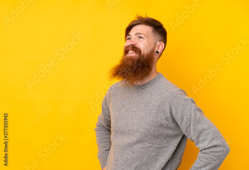 Redhead man with long beard over isolated yellow background posing with arms at hip and smiling