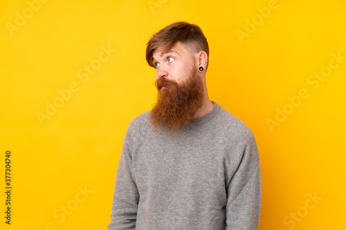 Redhead man with long beard over isolated yellow background making doubts gesture looking side