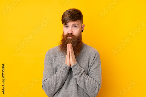 Redhead man with long beard over isolated yellow background keeps palm together. Person asks for something