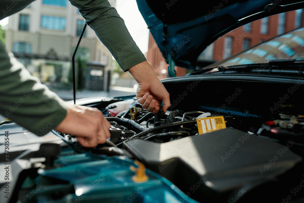 Close up of hands of young man examining broken down car engine, standing near his car with open hood on the city street