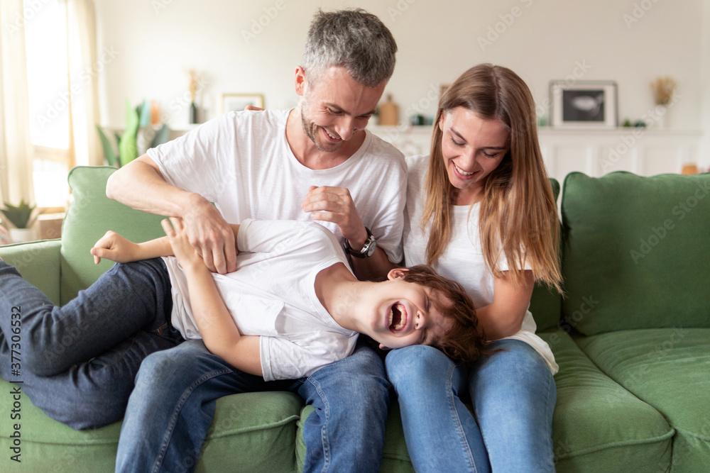 Happy young dad having fun tickling cute kid son relaxing on sofa together his wife. Modern carefree family laughing having fun with overjoyed kid on weekend.