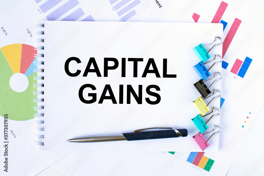 Notepad with text Capital Gains, paper clips, blue pen on financial diagrams Notepad with text Capital Gains, paper clips, blue pen on financial diagrams