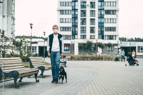 A teen boy walks on the street with his poodle dog. Coronavirus pandemic. A boy in a medical mask is observing social distancing measures.