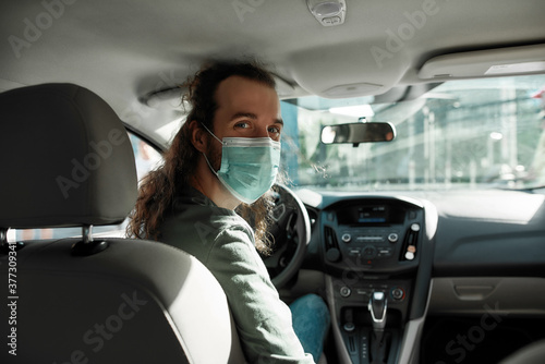 Man taxi driver talking to a passenger while steering the car during coronavirus pandemic wearing sterile medical mask, Social distance and health care concept