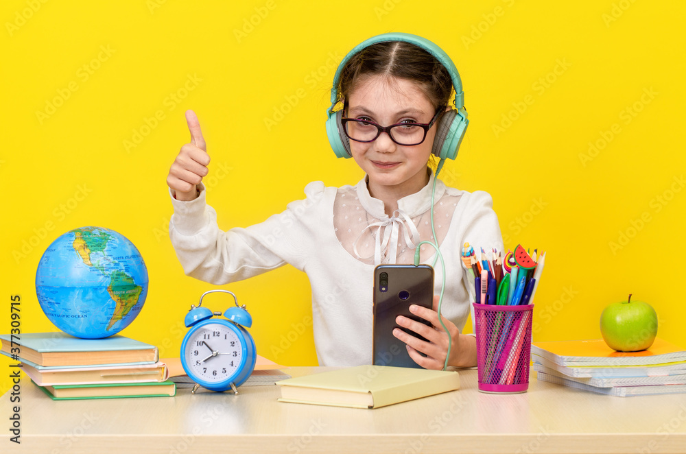 Back to school and happy time. Cute industrious child is sitting at a desk indoors. Kid is learning in class on yellow background. Happy smiling little girl with thumb up Back to school and happy time. Cute industrious child is sitting at a desk indoors. Kid is learning in class on yellow background. Happy smiling little girl with thumb up