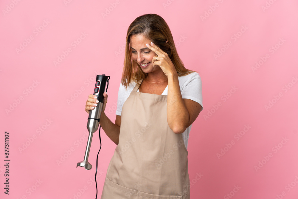 Middle age woman using hand blender isolated on pink background laughing