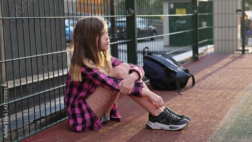 little / young lonely sad school girl in a pink plaid shirt with backpack sits alone on the street near the fence