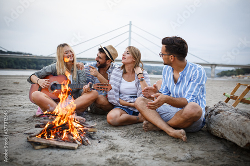 On a tranquil autumn evening, a group of friends gathers by the riverside sandy beach. They sit around a small campfire, under the starlit sky, enjoying their drinks, singing, and playing guitar.  © BalanceFormCreative