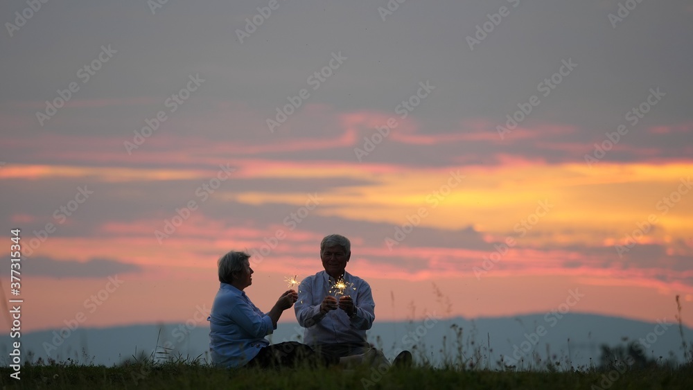 Fototapeta premium Couple of old people holding sparkling fire work and singing, sunset sky, marriage anniversary