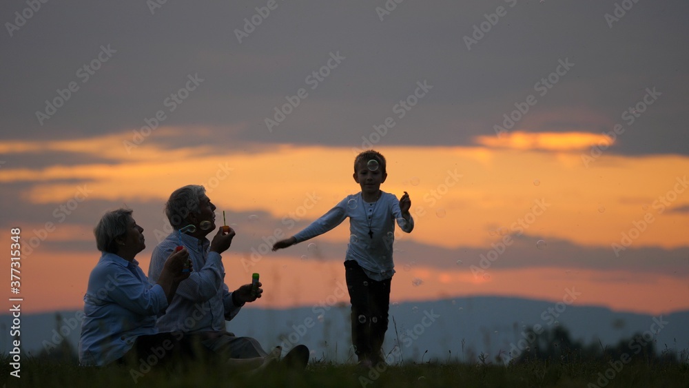 Grandparents blowing soap bubbles while grandchildren play to catch them at sunset in the nature