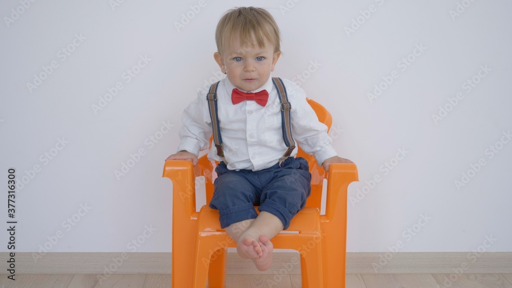 Funny blond hair baby child with blue eyes wear red bow and brace sit on chair, lovely boss Funny blond hair baby child with blue eyes wear red bow and brace sit on chair, lovely boss