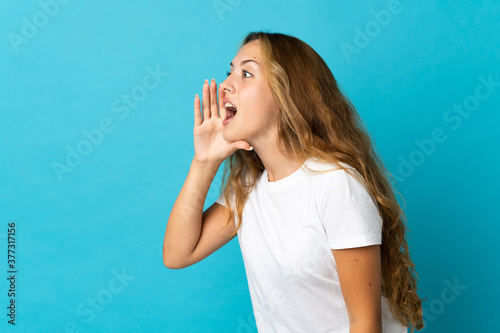 Young blonde woman isolated on blue background shouting with mouth wide open to the lateral