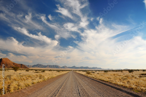 The Open Road. An unsealed scenic road in the Namibian desert, Africa, leading to a mountain range in the distance. Blue sky with wispy clouds.