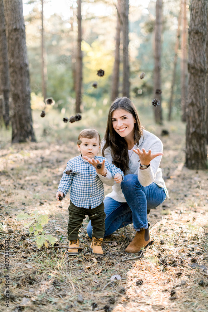 Outdoor portrait of smiling young joyful mom and little son, having fun and playing in beautiful pine autumn forest, throwing up fir cones and looking at camera Outdoor portrait of smiling young joyful mom and little son, having fun and playing in beautiful pine autumn forest, throwing up fir cones and looking at camera