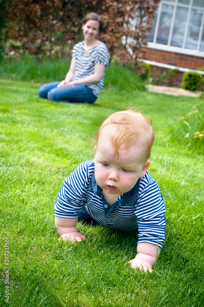 Proud mother watching young baby boy learning to crawl on the grass in the garden. Portrait format. Proud mother watching young baby boy learning to crawl on the grass in the garden. Portrait format.