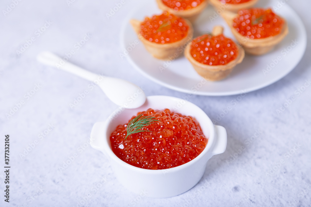 Red caviar (salmon caviar) in a white cup. In the background is a plate with tartlets and caviar. Selective focus, close up. Red caviar (salmon caviar) in a white cup. In the background is a plate with tartlets and caviar. Selective focus, close up.