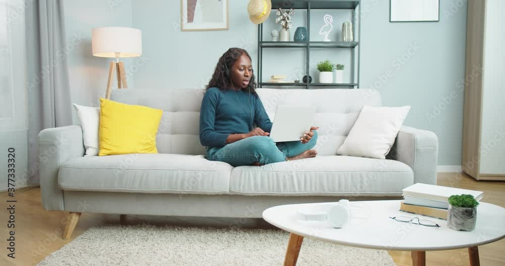 African American serious beautiful girl in headset having video conference while sitting on couch in room and typing on laptop. Pretty female texting on computer and talking on video at home indoors African American serious beautiful girl in headset having video conference while sitting on couch in room and typing on laptop. Pretty female texting on computer and talking on video at home indoors