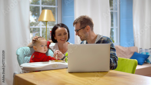 Wallpaper Mural Happy young couple feeding baby boy in highchair and using laptop at home Torontodigital.ca