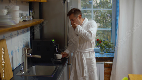 Young man in bathrobe making coffee and yawning in kitchen early in morning
