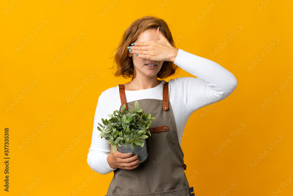 Young Georgian woman holding a plant isolated on yellow background covering eyes by hands. Do not want to see something Young Georgian woman holding a plant isolated on yellow background covering eyes by hands. Do not want to see something