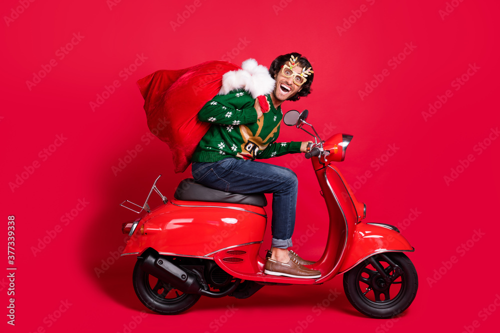 Profile side view portrait of attractive childish glad cheerful guy riding moped delivering gifts sack St Nicholas winter sale tradition isolated bright vivid shine vibrant red color background Profile side view portrait of attractive childish glad cheerful guy riding moped delivering gifts sack St Nicholas winter sale tradition isolated bright vivid shine vibrant red color background