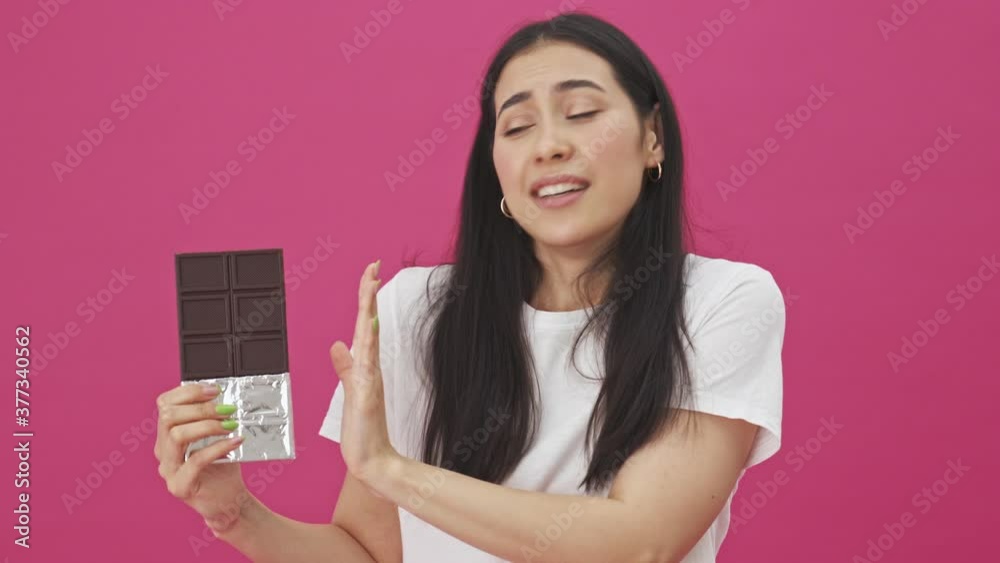 An attractive young woman is holding a chocolate bar while refusing of it standing isolated over a pink background An attractive young woman is holding a chocolate bar while refusing of it standing isolated over a pink background