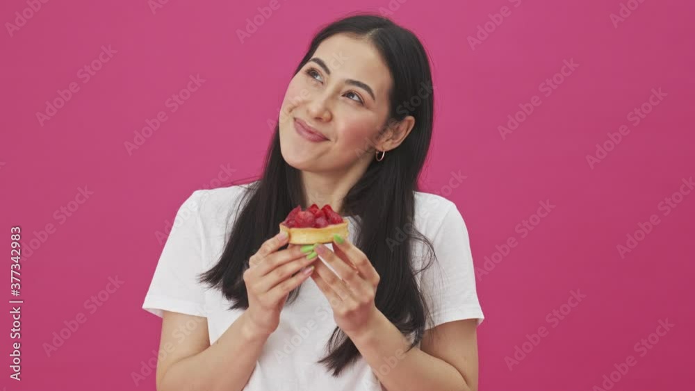 A smiling young woman is holding a strawberry cake while standing isolated over a pink background A smiling young woman is holding a strawberry cake while standing isolated over a pink background