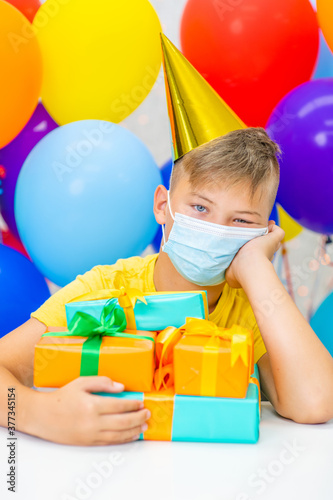 Unhappy boy wearing party cap and protective face mask sits at home with gift boxes during the coronavirus epidemic