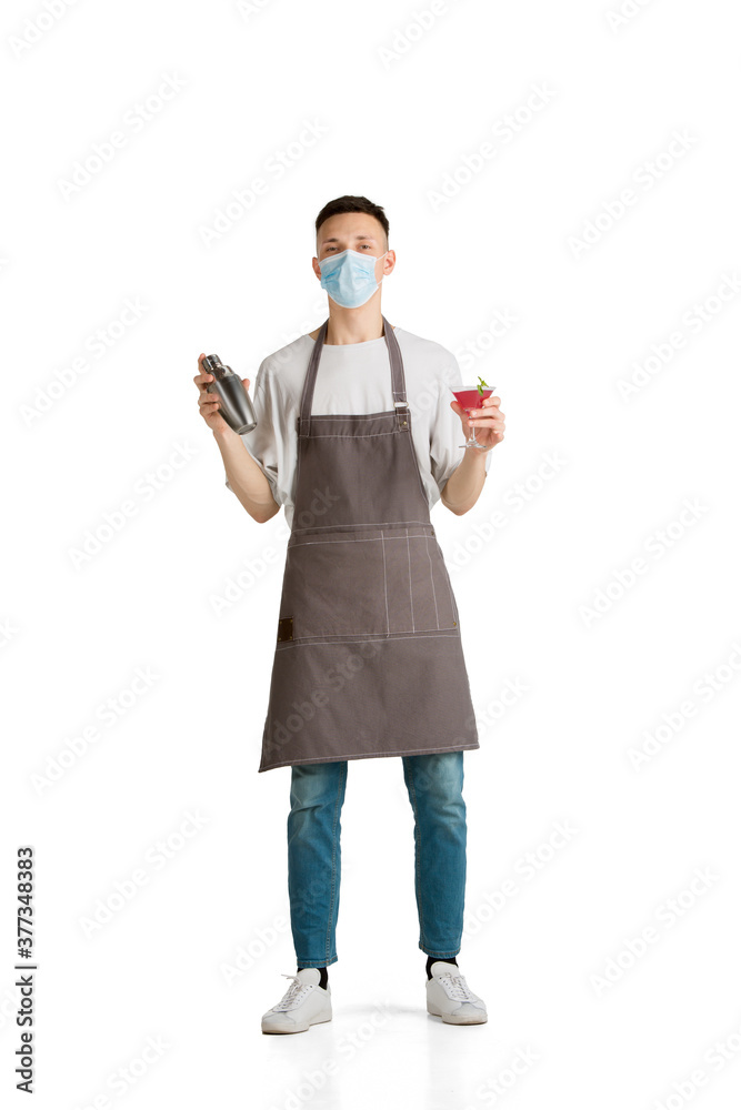 Shaker and drink. Portrait of a young male caucasian barista or bartender in brown apron. Studio white background, copyspace. Working in protective face mask. Professional occupation, drink, service. Shaker and drink. Portrait of a young male caucasian barista or bartender in brown apron. Studio white background, copyspace. Working in protective face mask. Professional occupation, drink, service.