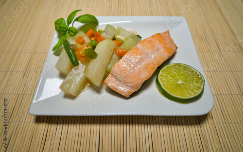 Gebratener Lachs mit Kohlrabi Gemüse, Karotten, Bohnen und einer Scheibe Limette auf einem weißen Teller serviert