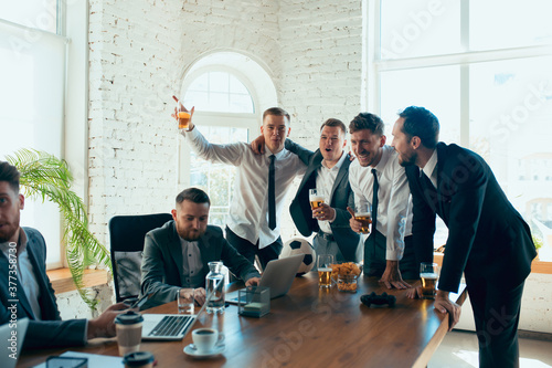 Watching football, drinking beer, cheering. Happy carefree colleagues having fun in office while their co-workers working hard and highly concentrated. Concept of fun, resting, professional occupation photo